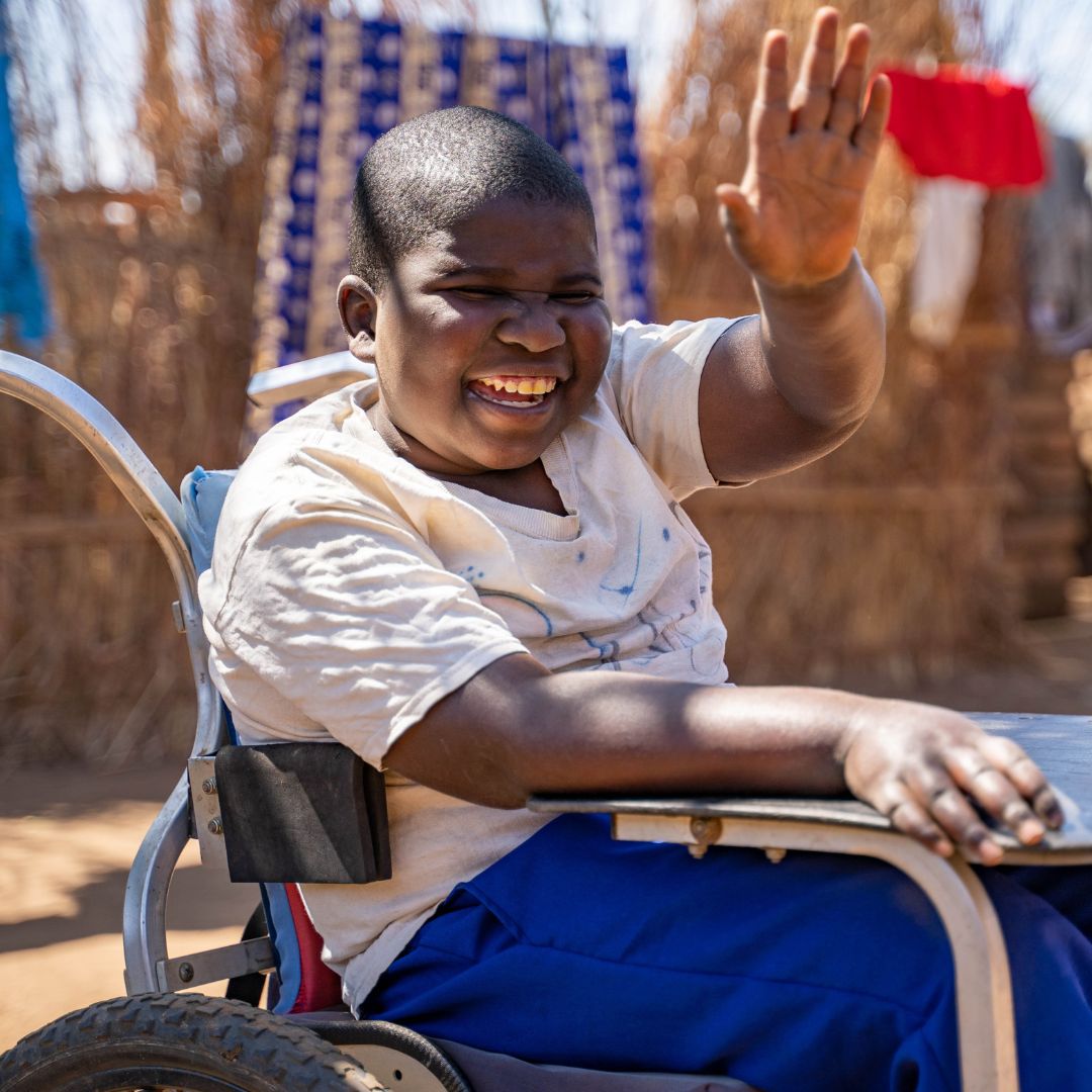 Schoolboy Enock is outside using his wheelchair while waving and smiling.